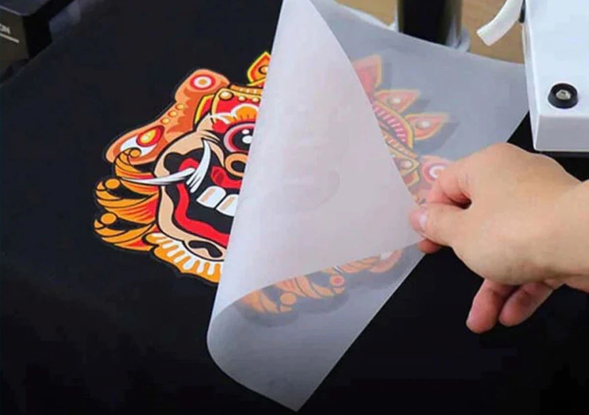 Hand peeling a DTF transfer film from a vibrant multicolor design on a black t-shirt